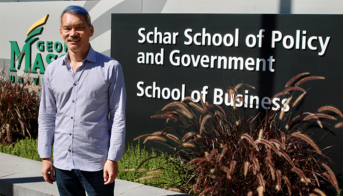 A man in a blue-gray shirt stands in front of the Mason Square Schar School sign.