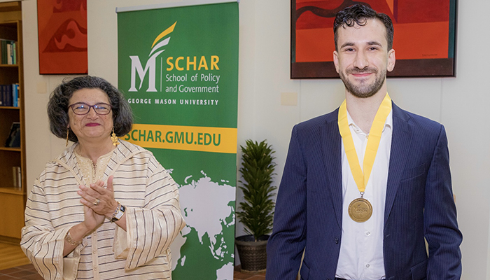 A man in a dark suit with a gold medal around his neck smiles as a woman applauds.