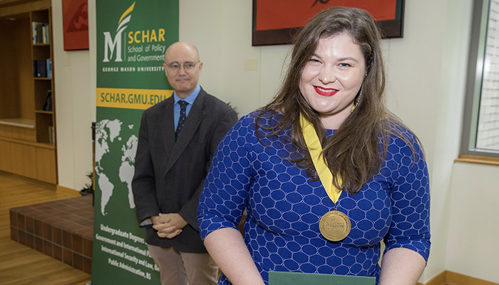 A woman in a blue dress holds a green certificate and wears a gold medal as a man in a dark jacket smiles in the background.