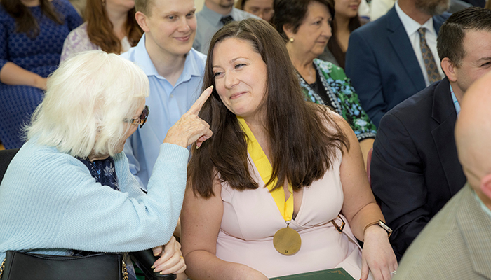 A woman in a pink dress wearing a gold medal smiles as a woman in white hair points a proud finger at her.