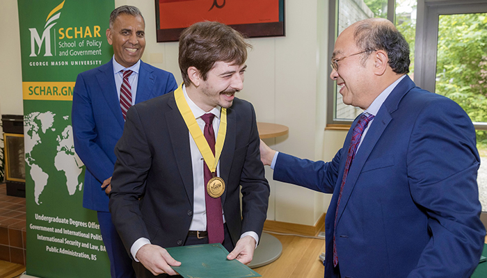 A man in a dark suit with a gold medal is congratulated by a man in glasses as a man in a blue suit smiles at the camera.