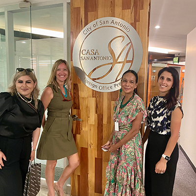 Four women stand in a wood paneled foyer in front of an official sign.