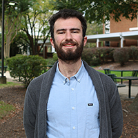 Colin McAulay stands outside on the George Mason University Fairfax Campus while wearing a gray sweater and blue collared shirt.