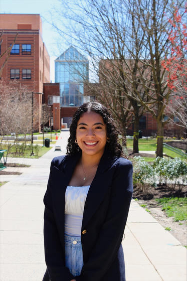 A young woman with dark hair wearing a dark blazer smiles stands on a walkway on George Mason University’s campus and smiles at the camera.