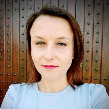 Ludmila Oleinic in a blue top and pink lipstick stands in front of a brown wall.