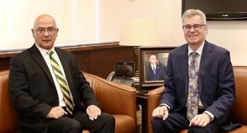 Two men in suits and ties sit on brown leather chairs looking at the camera.