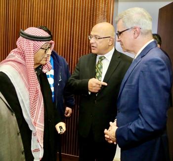 A man in an Arab headscarf and two men in suits and ties stand in front of a brown wall.