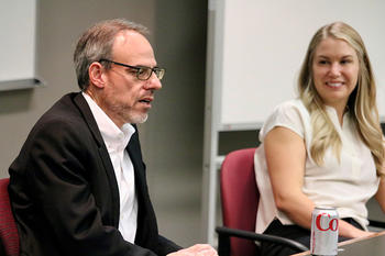 A woman in a white blouse smiles as a man in a dark jacket and white shirt in glasses speaks.