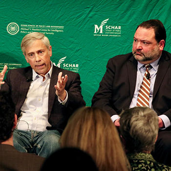 A man in a suit without a tie gestures with his hands as another man in a dark suit watches.