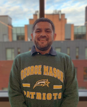 A bearded man in a George Mason University sweatshirt stands in front of a window.