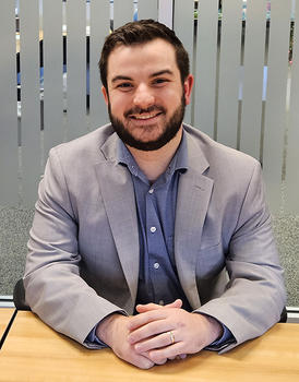 A young man with a beard sits at a conference room table and smiles at the camera.