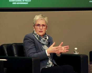 A woman in red eyeglasses, a gray jacket, and a gray and white scarf gestures with her hands.