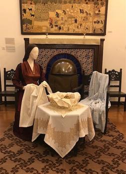 A female mannequin dressed in traditional Greek clothing sits at a table in front of an ornate fireplace.