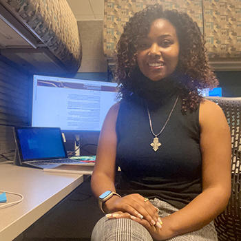 A young woman wearing a black sleeveless top sits in front of a computer in a cubicle.